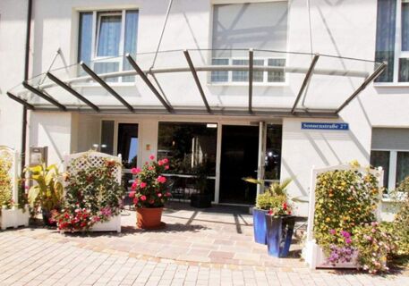 Entrance of Apartments Aschheim with glass canopy, potted flowers, plants, white lattice fences, and sliding glass doors