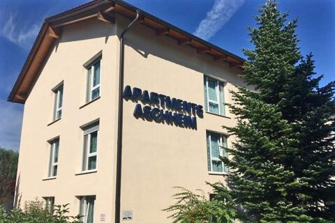 A beige three-story building with multiple windows and the name Apartments Aschheim on the wall under a blue sky and next to evergreen trees
