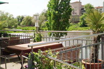 Hotel-Caf&eacute; 3 Kronen'in yazlık terasında masalar, ahşap banklar ve arka planda nehir ve yeşil ağa&ccedil;lar manzarası.