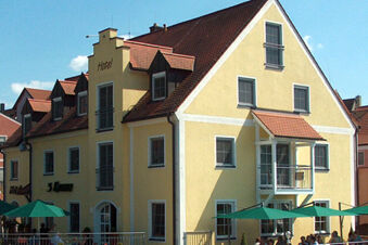 Hotel-Caf&eacute; 3 Kronen'in kırmızı &ccedil;atılı sarı binasının &ouml;n cephesi ve g&uuml;neşli bir g&uuml;nde yeşil şemsiyeler altında a&ccedil;ık havada oturma yerleri.