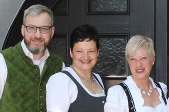 Bir girişin &ouml;n&uuml;nde geleneksel kıyafetler i&ccedil;inde dost&ccedil;a g&uuml;l&uuml;mseyen &uuml;&ccedil; kişi, sıcak misafirperverliği ile Hotel-Caf&eacute; 3 Kronen'i temsil ediyor.