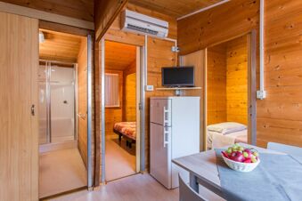 Interior of a wooden cabin with a refrigerator, wall-mounted TV, air conditioner, two beds, and a fruit basket at Marina Romea Easy Camping Village