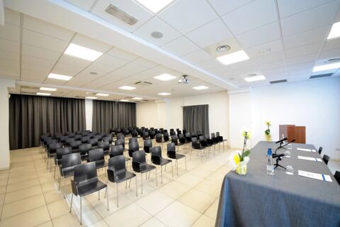 Conference room at B&B Hotel Mantova Cristallo with rows of black chairs, a table with microphones, and tiled flooring