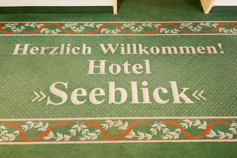 Green carpet with white text "Herzlich Willkommen! Hotel Seeblick" surrounded by decorative leaf patterns for Land-gut-Hotel Seeblick