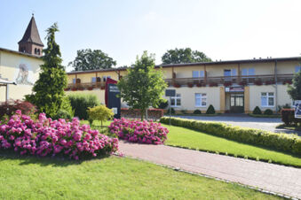 Land-gut-Hotel Seeblick building with two floors, pink flower bushes, green trees, and a church steeple in the background