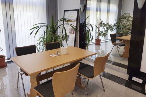 Simple dining area with a wooden table, four chairs with dark cushions, large plants, and a Land-gut-Hotel Seeblick sign