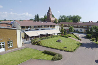 Land-gut-Hotel Seeblick with a lawn, paved paths, outdoor seating under awnings, and a church with a tower in background