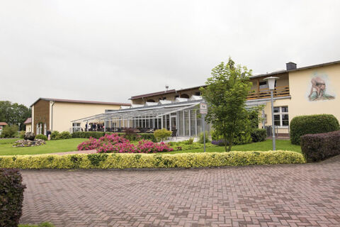Land-gut-Hotel Seeblick building with glass patio, green lawn, pink flowers, trees, and a brick walkway under cloudy sky