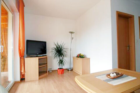 Living room at Land-gut-Hotel Seeblick with wooden furniture, a TV on a stand, potted plant, floor lamp, and sliding glass door with orange curtains