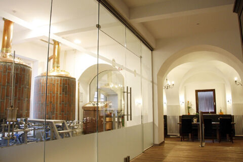Interior view of Land-gut-Hotel Seeblick showing copper brewing tanks behind glass and a dining area with white walls and wooden floor