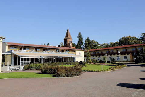 Land-gut-Hotel Seeblick building with red roof, covered terrace, green garden, brick driveway, and church tower in background