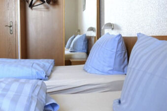 Enkelt inrett sovrum med bl&aring; s&auml;ngkl&auml;der och tr&auml;m&ouml;bler i Gasthof Pension R&ouml;per-Bolte, inklusive v&auml;ggspegel och garderob.