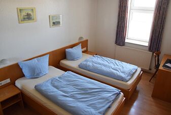 Enkelt inrett hotellrum med tv&aring; enkels&auml;ngar, bl&aring;tt s&auml;nglinne och f&ouml;nster i Gasthof Pension R&ouml;per-Bolte.