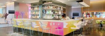 Modern bar area with colorful gradient counter, transparent and white chairs, and shelves stocked with bottles at Nhow Milano