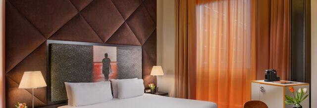 Hotel room at Nhow Milano with a white bed, two lamps, brown padded wall panels, artwork, and sheer orange curtains on the window