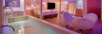 Indoor pool next to a bedroom with a bed, TV, vanity desk, large mirror, and two white curved chairs at Nhow Milano
