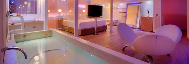 Indoor pool next to a bedroom with a bed, TV, vanity desk, large mirror, and two white curved chairs at Nhow Milano