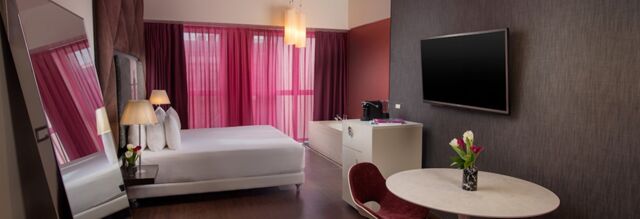 Modern hotel room at Nhow Milano featuring a large bed, purple curtains, a round table with flowers, and a flat-screen TV