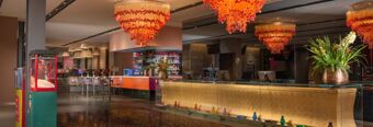 Modern hotel lobby with a golden reception desk, orange chandeliers, bar area, popcorn machine, and floral decorations at Nhow Milano