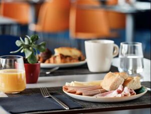 A breakfast plate with sliced ham, cheese, bread rolls, a glass of orange juice, a white coffee cup, and water at Holiday Inn Express Milan-Malpensa Airport