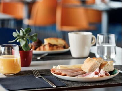 A breakfast plate with sliced ham, cheese, bread rolls, a glass of orange juice, a white coffee cup, and water at Holiday Inn Express Milan-Malpensa Airport