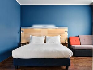 A neatly made bed with white linens and pillows against a blue wall next to a sofa in Holiday Inn Express Milan-Malpensa Airport