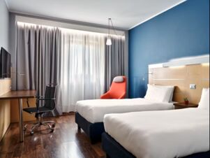 Hotel room at Holiday Inn Express Milan-Malpensa Airport with two single beds, blue accent wall, desk, office chair, and bright window coverings