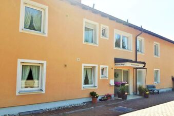 Fassade von Hotel Pension Heigl mit gelb-orangener Au&szlig;enwand, mehreren Fenstern und einem Eingangsbereich mit Pflanzen und Sitzbank.