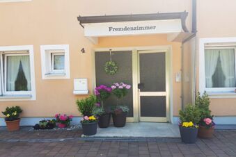 Eingangsbereich mit Haust&uuml;r, Blumendekoration und dem Schild Fremdenzimmer des Hotel Pension Heigl in hellgelbem Geb&auml;ude.