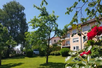 Gr&uuml;ne Gartenanlage mit B&auml;umen und Blumen vor der Fassade des Hotel Pension Heigl an einem sonnigen Tag.