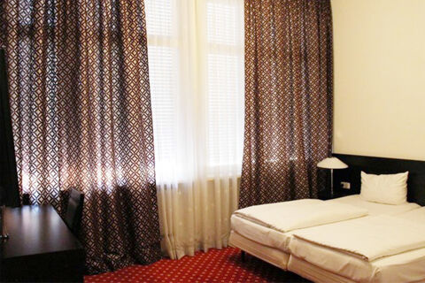 Světl&yacute; pokoj v hotelu Aariana s manželskou postel&iacute;, nočn&iacute; lampičkou, psac&iacute;m stolem a vzorovan&yacute;mi z&aacute;věsy na velk&yacute;ch oknech.