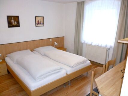A simple hotel room at Gasthaus Berger with two single beds joined together, wooden floors, desk, chairs, and window with curtains