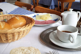 A breakfast setting at Gasthaus Berger with a basket of rolls, sliced cheese, cold cuts, a cup of coffee, and a white teapot on a table