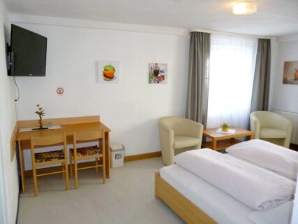 Bright hotel room at Gasthaus Berger with a double bed, two armchairs, a wooden desk, and a wall-mounted TV