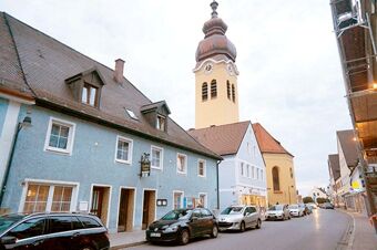 Street view of the blue Gasthaus Berger building with parked cars and a church tower in the background under a cloudy sky