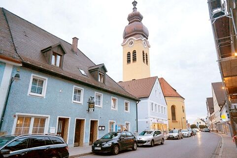 Street view of the blue Gasthaus Berger building with parked cars and a church tower in the background under a cloudy sky