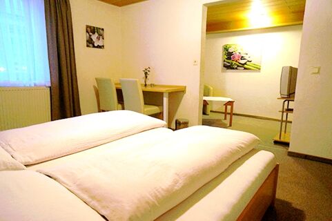 A bright hotel room at Gasthaus Berger with two beds, a wooden table with two chairs, a window with curtains, and a TV in an adjacent area