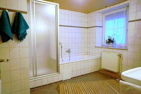 Bathroom in Gasthaus Berger featuring a bathtub, a shower with frosted glass doors, towels hanging on hooks, and a window with a flower arrangement