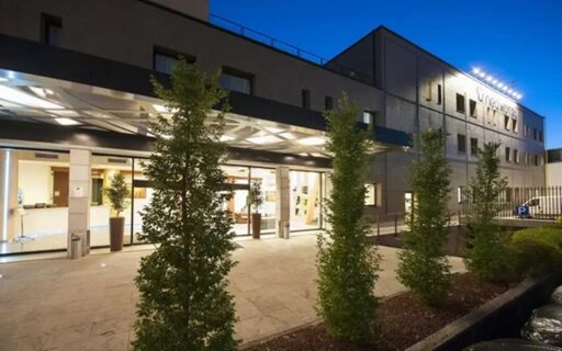 NEO Hotel exterior at night showing the entrance with glass doors and several tall green shrubs in front