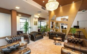 A hotel lobby at NEO Hotel featuring several leather armchairs and sofas, wooden tables, plants, and a large chandelier