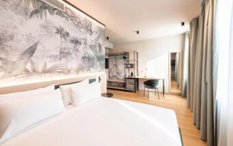 Bright hotel room at NEO Hotel featuring a large bed, a desk with a chair, wardrobe, and a window with curtains