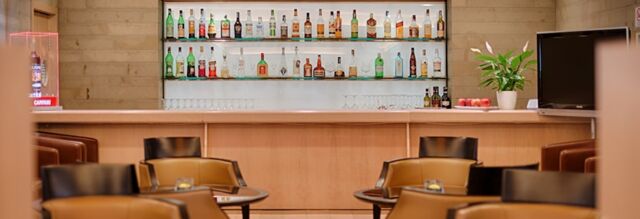 Bar area with shelves of various liquor bottles, beige wood paneling, black and brown chairs, and a TV at NH Hotel Linate