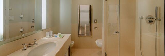 Bathroom in NH Hotel Linate with a large mirror, sink, countertop toiletries, towel rack, toilet, and glass shower enclosure