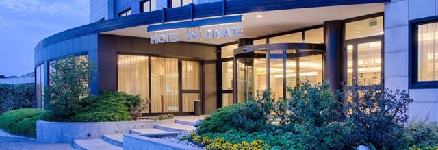 Exterior view of NH Hotel Linate entrance at dusk with revolving glass door, illuminated windows, and landscaped greenery surrounding steps