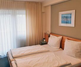 Helles Hotelzimmer im Hotel Frisch mit zwei Einzelbetten, wei&szlig;en Bettw&auml;sche, einem Nachttisch und gro&szlig;em Fenster mit Gardinen.