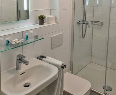 Modernes, sauberes Badezimmer mit Dusche, Waschbecken und WC im Hotel Frisch, ausgestattet mit wei&szlig;en Fliesen und frischen Handt&uuml;chern.