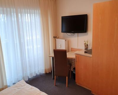 Hell eingerichtetes Zimmer im Hotel Frisch mit einem Schreibtisch, braunem Stuhl, Wandfernseher und gro&szlig;em Fenster mit Gardinen.
