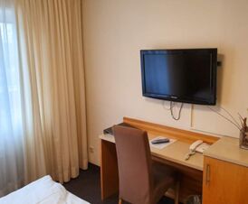 Ein schlicht eingerichteter Zimmerbereich im Hotel Frisch mit Tisch, Stuhl, Telefon und an der Wand montiertem Fernseher vor Gardinen.