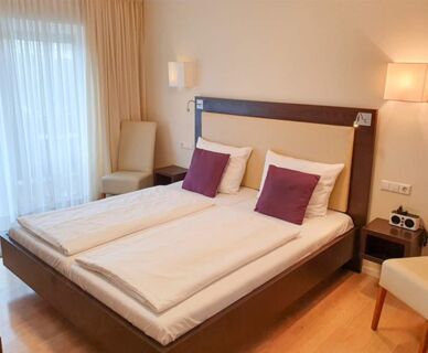 Modern eingerichtetes Schlafzimmer im Hotel Frisch mit zwei Einzelbetten, wei&szlig;en Bettw&auml;sche und lila Kissen sowie Tageslicht durch gro&szlig;e Fenster.