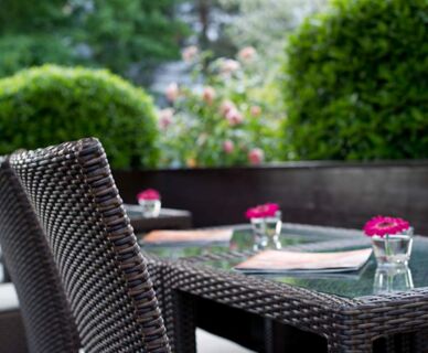 Bequeme Rattan-St&uuml;hle an einem Gartentisch mit kleinen Gl&auml;sern und pinken Blumen im Au&szlig;enbereich des Hotel Frisch.